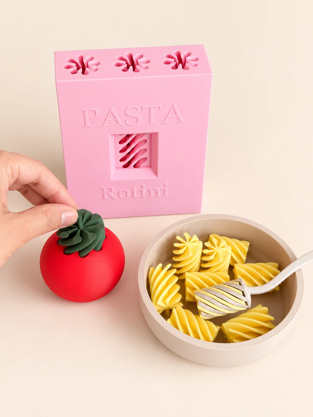Set de pasta con caja rosada, tomate con espiral verde, plato con fideos amarillos y tenedor, sobre fondo crema
